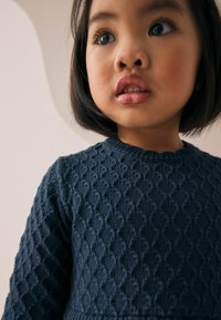 Pull en maille bleu marine foncé avec un motif de diamants texturé, encolure ronde et poignets côtelés. Le visage du modèle est partiellement visible.