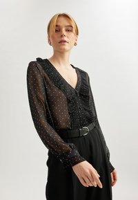 Sort gennemsigtig bluse med hvide stipletter, flæset halsudskæring og knaplukning. Lange ærmer med samlede manchetter, parret med et sort bælte.