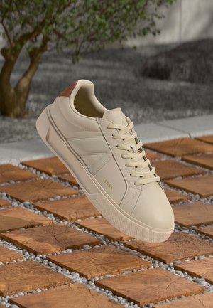 Krémově zbarvená teniska se šňůrkami a hnědým jazýčkem na patě, která levituje nad dřevěným blokem a štěrkovou cestou s rozostřeným pozadím stromu.