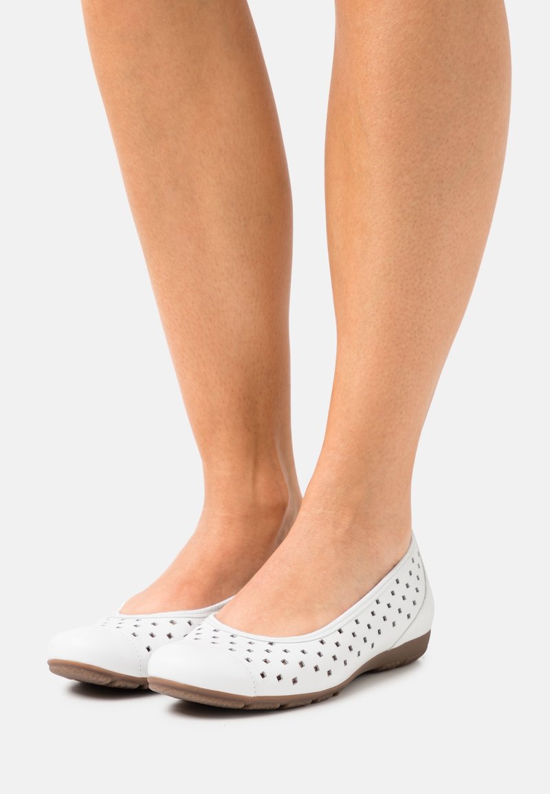 Pieds portant des chaussures plates blanches avec de petites perforations carrées et des semelles marron, sur un fond clair.
