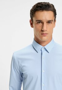 Camicia a righe di colore blu chiaro con maniche lunghe, colletto a punta e chiusura frontale con bottoni. Tessuto liscio con una sottile texture.