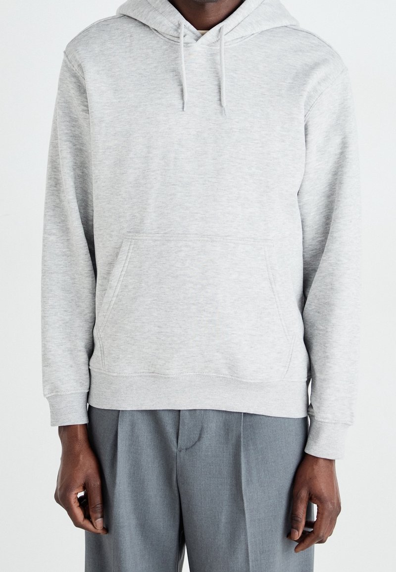 Homme portant un sweat à capuche gris clair avec poche frontale et pantalon gris, debout devant un fond blanc uni.