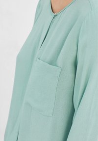 Helle türkisfarbene Bluse mit einer Tasche, weicher Textur, rundem Halsausschnitt und einem halben Reißverschluss. Mit lockeren, fließenden Ärmeln.