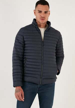 Marineblå pufferjakke med horisontalt quiltet struktur, lynlås lukning, høj krave og sidelommer. Båret over en cremet sweater og mørke jeans.