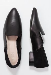 Svart läderplatta med spetsiga tår, öppna sidor och låg blockklack. Har en slät textur och minimalistisk design.