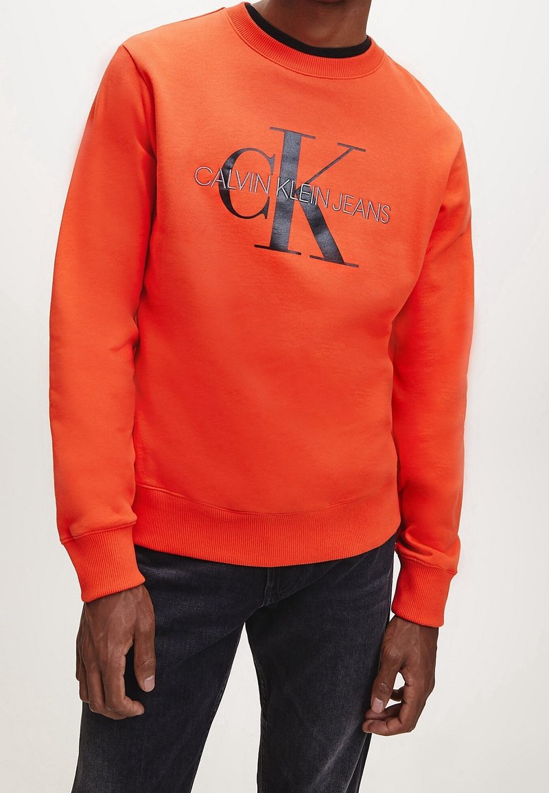Jasnopomarańczowa bluza z okrągłym dekoltem, z dużym czarnym logo Calvin Klein Jeans na przodzie. Ściągacze na mankietach i dole bluzy.