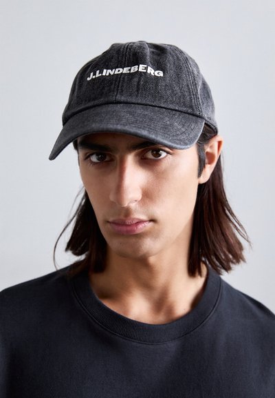 Căciulă din denim negru cu brâu curbat și logo alb "J.Lindeberg". Textură netedă și design structurat, purtată cu un hanorac negru.