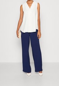 Haut blanc sans manches avec un col en V, associé à un pantalon bleu marine à jambes larges. Accessoires : un petit sac à main crème et des talons blancs.