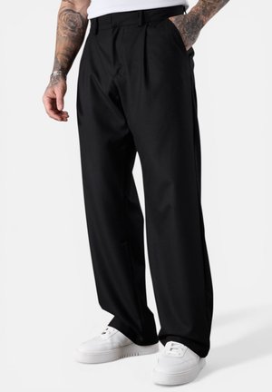 Man wearing black wide-leg trousers, white sneakers, and a white top, with tattooed arms and hands in pockets.