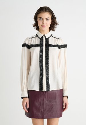 Blouse crème avec des accents et des détails en dentelle noire, dotée d'un devant boutonné et de manches longues, associée à une jupe en cuir bourgogne.