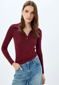 Burgundy pitkähiha polo paita, jossa on kaulus ja kolmen napin lista; yhdistetty korkeavyötäröisiin sinisiin farkkuihin; sileä rakenne.