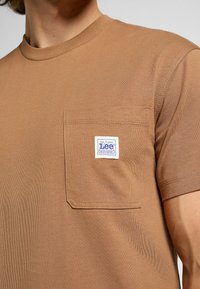 Brun bomulls-T-shirt med rund halsringning, korta ärmar och en vänster bröstficka som har en sytt etikett med "Lee." Slät textur.
