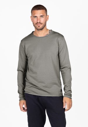 Camisa de manga larga en verde olivo, hecha de una tela suave con un cuello redondo y detalles de costura sutiles. Combinada con pantalones oscuros.