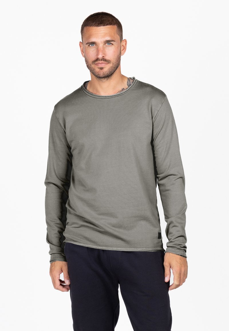 Langärmliges Shirt in Olivgrün, aus glattem Stoff mit rundem Halsausschnitt und feinen Nähten. Kombiniert mit dunklen Hosen.