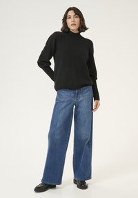 Pull en tricot noir avec un col montant et des manches bouffantes, associé à un jean en denim bleu à jambes larges. Des chaussures à plateforme noires complètent le look.