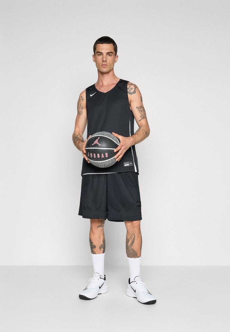 Uomo con tatuaggi in abbigliamento da basket nero che tiene un pallone da basket Jordan nero e grigio, indossando scarpe e calze Nike bianche.