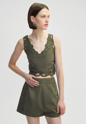 Olivově zelený crop top s krajkovým lemem a výřezovými detaily, kombinovaný s odpovídajícími šortkami s vysokým pasem, které mají podobné dekorativní prvky.