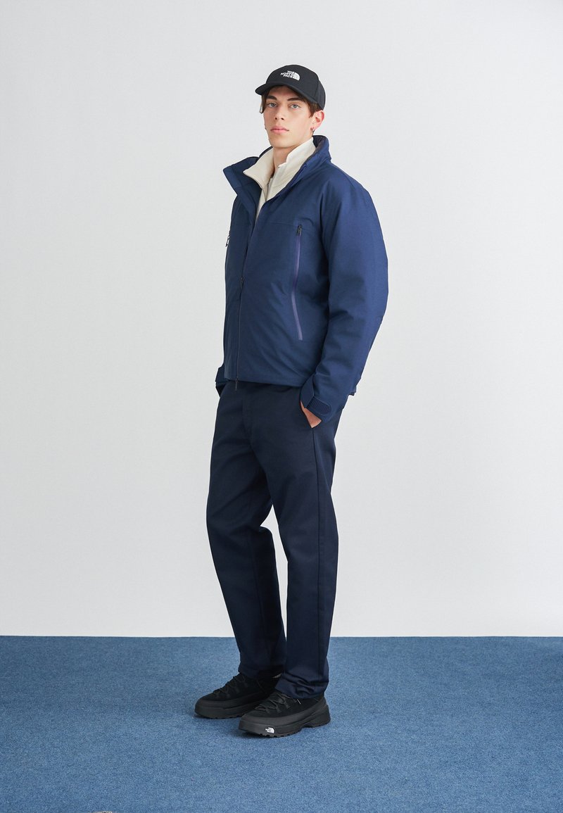 Veste isolée bleu marine avec un col haut, doublure intérieure légère et accents violets, portée avec un pantalon foncé et des chaussures noires.