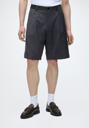 Mand iført mørkegrå plisserede shorts, hvide crew-sokker og mørkebrune læder loafers med kvaster, stående mod en ensfarvet baggrund.
