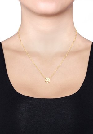Collier en or avec un pendentif de visage souriant simple, doté d'une chaîne délicate et d'une finition poli, porté sur un haut noir.