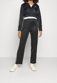 Person som bär en svart Adidas träningsjacka och byxor med vita ränder, kombinerat med vita sneakers och rosa strumpor, stående mot en enhetlig bakgrund.