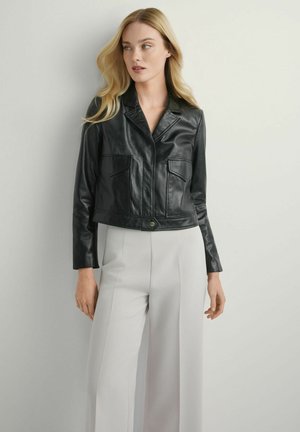 Kobieta o blond włosach, ubrana w czarną skórzaną kurtkę i jasnoszare, szerokie spodnie z wysokim stanem, stoi na tle jednolitej białej ściany.