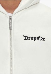 Biała bluza z kapturem na zamek, wykonana z miękkiego materiału. Posiada czarne haftowane logo "Dropsize" oraz srebrny zamek. Model z kapturem.