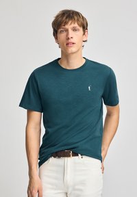 T-shirt azzurro a maniche corte realizzato in un morbido tessuto, con collo tondo e un piccolo logo bianco sul lato sinistro del petto. Indossato con pantaloni bianchi.