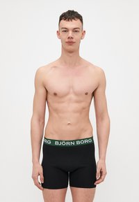 Black boxer shorts with a green waistband featuring "BJÖRN BORG" text. Smooth fabric, fitted design, with stitching details.