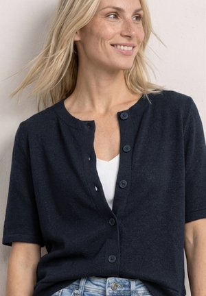 Blonde Frau lächelt, trägt eine marineblaue kurzärmelige, geknöpfte Strickjacke über einem weißen Top mit hellblauen Jeans vor neutralem Hintergrund.