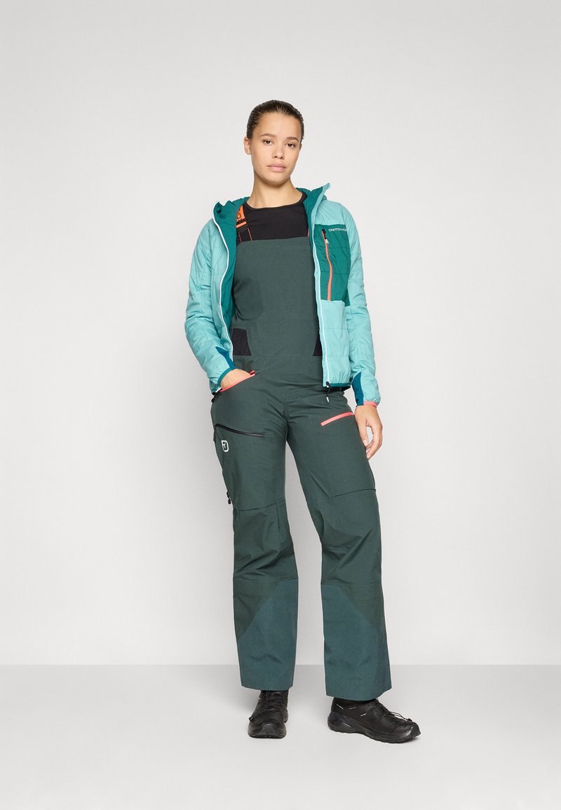 Grüne Latzhose mit schwarzen Akzenten, eine türkisfarbene leichte Jacke und schwarze Stiefel. Verfügt über Taschen und Reißverschlüsse, entworfen für den Einsatz im Freien.
