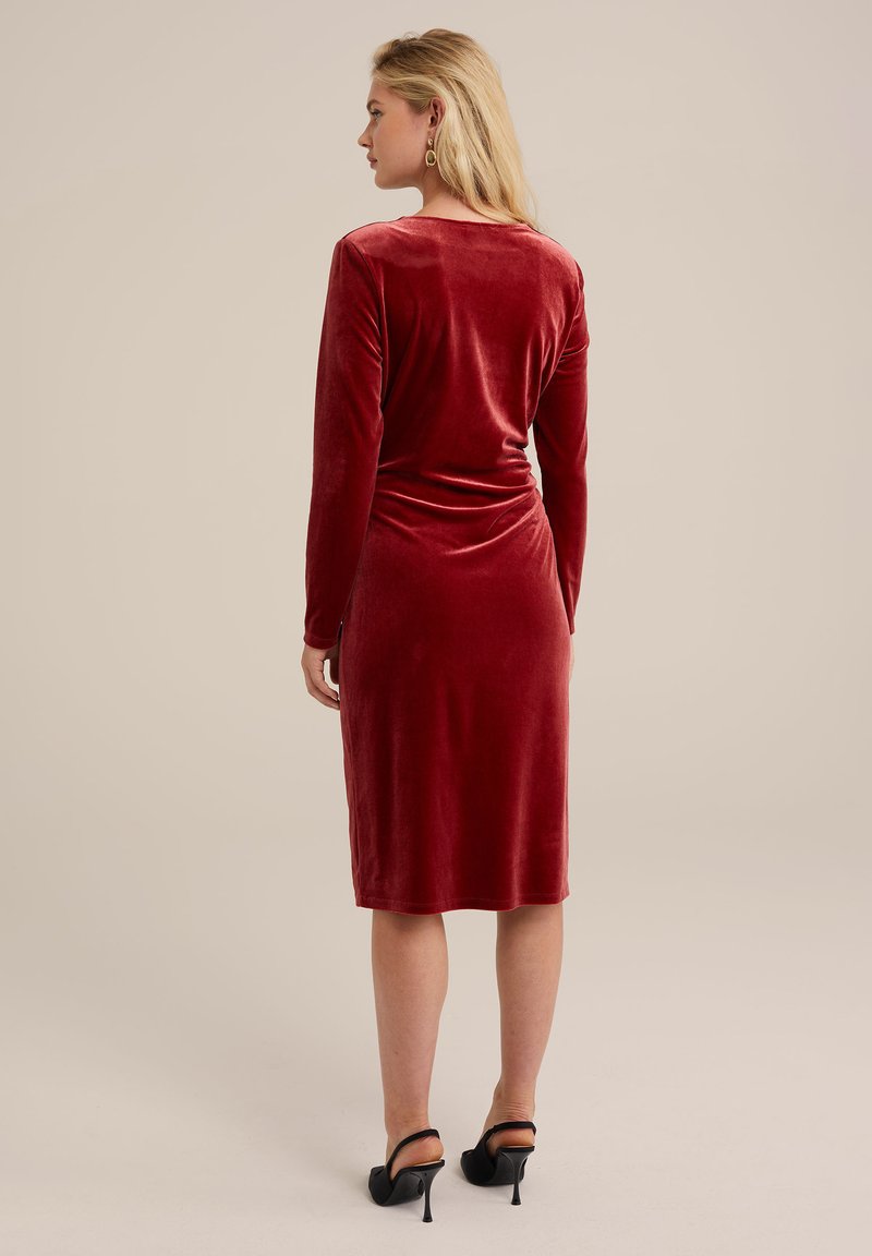 Czerwona aksamitna sukienka z długimi rękawami, do kolan, dopasowany krój, gładka tekstura, z marszczeniem z tyłu. Noszona z czarnymi szpilkami.