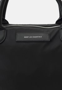 Svart tote-väska i nylon med läderdetaljer, utrustad med en dragkedja upptill och en etikett med texten "WANT LES ESSENTIELS." Slät textur.