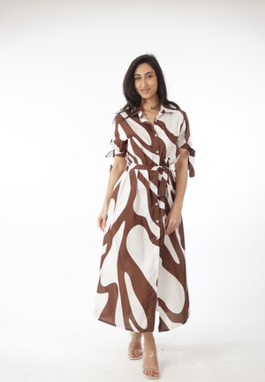 Kvinde iført en brun og hvid midi-kjole med knapper, abstrakt mønster, korte ærmer og bånd i taljen, stående foran en hvid baggrund.