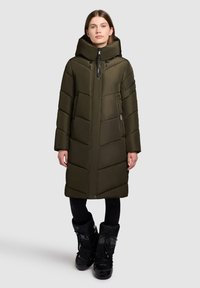 Olijfgroene pufferjas met hoge kraag, ritssluiting en zijzakken. Voorzien van gequiltte panelen en een gestroomlijnd ontwerp. Zwarte winterlaarzen gedragen.