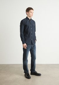 Donkerblauw overhemd met knopen in een zachte stof, gekenmerkt door een subtiel logo op de borst, gecombineerd met blauwe jeans en zwarte gestructureerde schoenen.