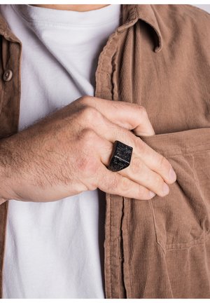 Black rectangular ring with textured detailing, worn on a hand holding a brown corduroy shirt pocket, over a white T-shirt.