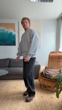 Sweatshirt cinza com corte descontraído, jeans escuros e sapatos castanhos. O fundo apresenta um sofá cinza, uma cadeira tecida e arte de parede abstrata.
