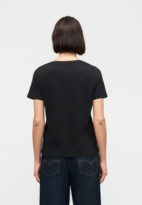 Άτομο με κοντά σκούρα μαλλιά, φοράει απλό μαύρο κοντομάνικο μπλουζάκι και σκούρο μπλε τζιν, με την πλάτη προς τα εμπρός σε μονόχρωμο φόντο.