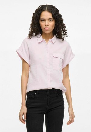 Světle růžová košile s krátkým rukávem a límečkem, s přední kapsou a texturovanou látkou. V kombinaci s černými džíny.