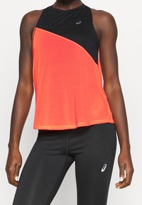 Sportbekleidung mit ärmellosem orangen und schwarzen Oberteil sowie schwarzen Leggings mit Logos, getragen von einer dunkelhäutigen Person, die mit entspannten Armen dasteht.