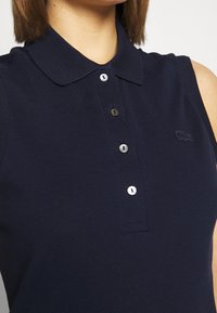 Mörkblå ärmlös polo med krage, tre-knappars knappslå, strukturerad tyg och en liten logotyp på bröstet.