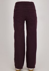 Vrouw in losse donkerpaarse broek met achterzakken, staand tegen een effen lichte achtergrond.