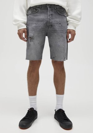 Homme portant un short en denim gris usé, des chaussettes blanches, des baskets noires et un sweat-shirt blanc, debout devant un fond uni.