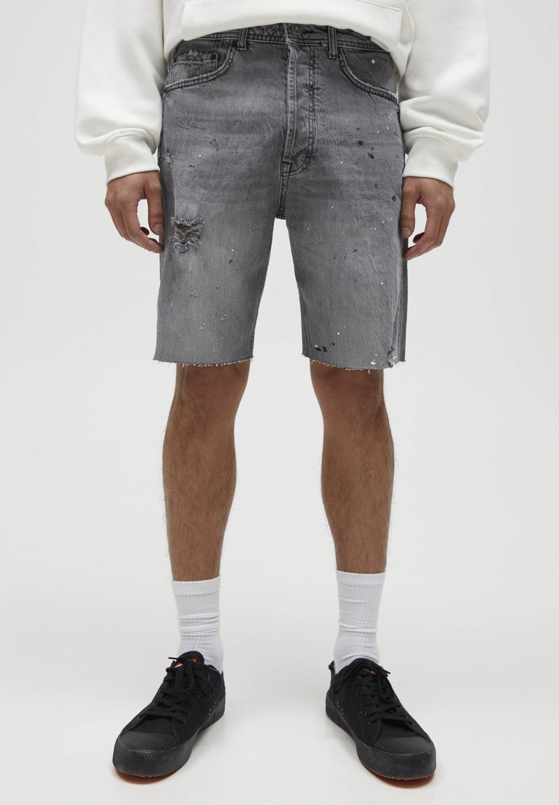 Homme portant un short en denim gris usé, des chaussettes blanches, des baskets noires et un sweat-shirt blanc, debout devant un fond uni.