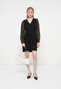 Vestido preto com mangas transparentes de bolinhas, decote em V e cintura com cordão. A modelo segura uma bolsa creme, combinada com sapatos pretos de bico fino.