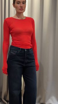 Žena v přiléhavé červené košili s dlouhým rukávem a tmavých džínách s vysokým pasem stojí před béžovými závěsy.