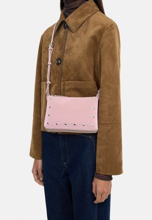 Osoba ubrana w brązową zamszową kurtkę i ciemnoniebieskie dżinsy, z małą różową torebką na ramię ozdobioną srebrnymi ćwiekami.