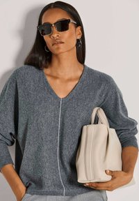 Frau mit schwarzen quadratischen Sonnenbrillen und grauem Pullover, die mit einer Hand eine beigefarbene Handtasche gegen einen hellen Hintergrund hält.