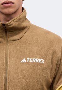 Κοντινό πλάνο ατόμου που φοράει καφέ fleece μπουφάν με ψηλό γιακά και λευκό λογότυπο Terrex στο στήθος.